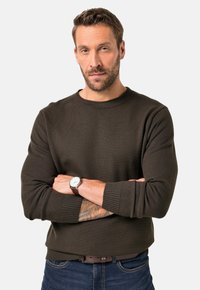 Dunkelbrauner Strickpullover mit Rundhalsausschnitt, gerippten Bündchen und Saum. Das Modell trägt eine Uhr und steht mit verschränkten Armen, um die Textur zu zeigen.