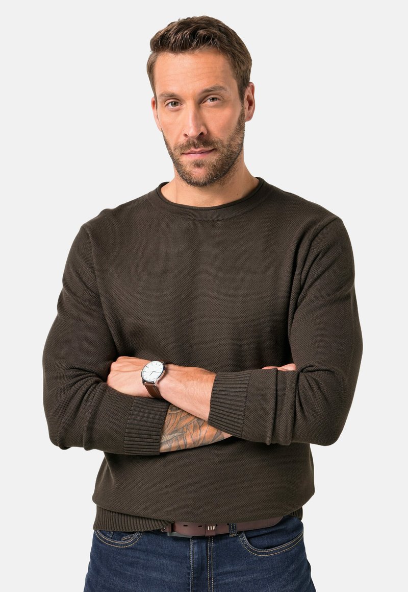 Dunkelbrauner Strickpullover mit Rundhalsausschnitt, gerippten Bündchen und Saum. Das Modell trägt eine Uhr und steht mit verschränkten Armen, um die Textur zu zeigen.