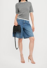 Suéter gris de manga corta con ribete blanco, pantalones cortos de mezclilla azul, bolso negro con flecos y sandalias de tacón beige. Diseño sencillo y atuendo casual.