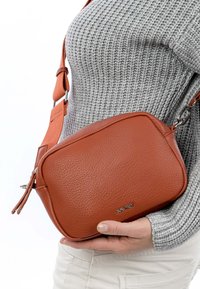 Persoon draagt een grijze gebreide trui en witte broek en houdt een kleine bruine leren crossbodytas met een verstelbare riem vast.