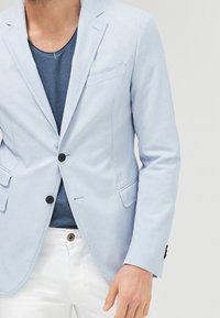 Světle modrý bavlněný blazer se dvěma předními kapsami, zapínáním na jeden knoflík, klopami a slim fit, kombinovaný s námořnicky modrým tričkem a bílými kalhotami.