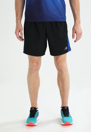 Mężczyzna ubrany w czarne sportowe spodenki z niebieskimi bocznymi panelami oraz turkusowe buty do biegania z pomarańczowymi podeszwami, stojący na białej podłodze.