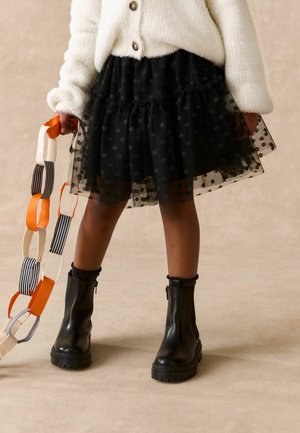 Kind trägt schwarzen Tüllrock mit Punkten, weißen flauschigen, geknöpften Pullover, schwarze Stiefel, hält eine orange-schwarz gestreifte Papierkette.