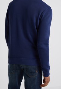 Mörkblå sweatshirt med ribbade ärmslut och nederkant, tillverkad av ett slätt, texturerat tyg, ihop med mörka jeans med subtil sömnad vid bakfickorna.
