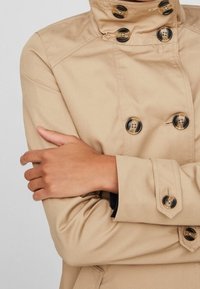 Beige trenchcoat met hoge kraag, dubbele knopen en grote donkere knopen. De stof lijkt glad met zichtbare stikdetails.