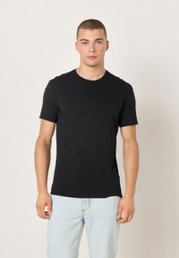 Crna pamučna majica s okruglim izrezom, kratkim rukavima i prilagođenim krojem. Nošena s svijetloplavim trapericama, prikazana na neutralnoj pozadini.