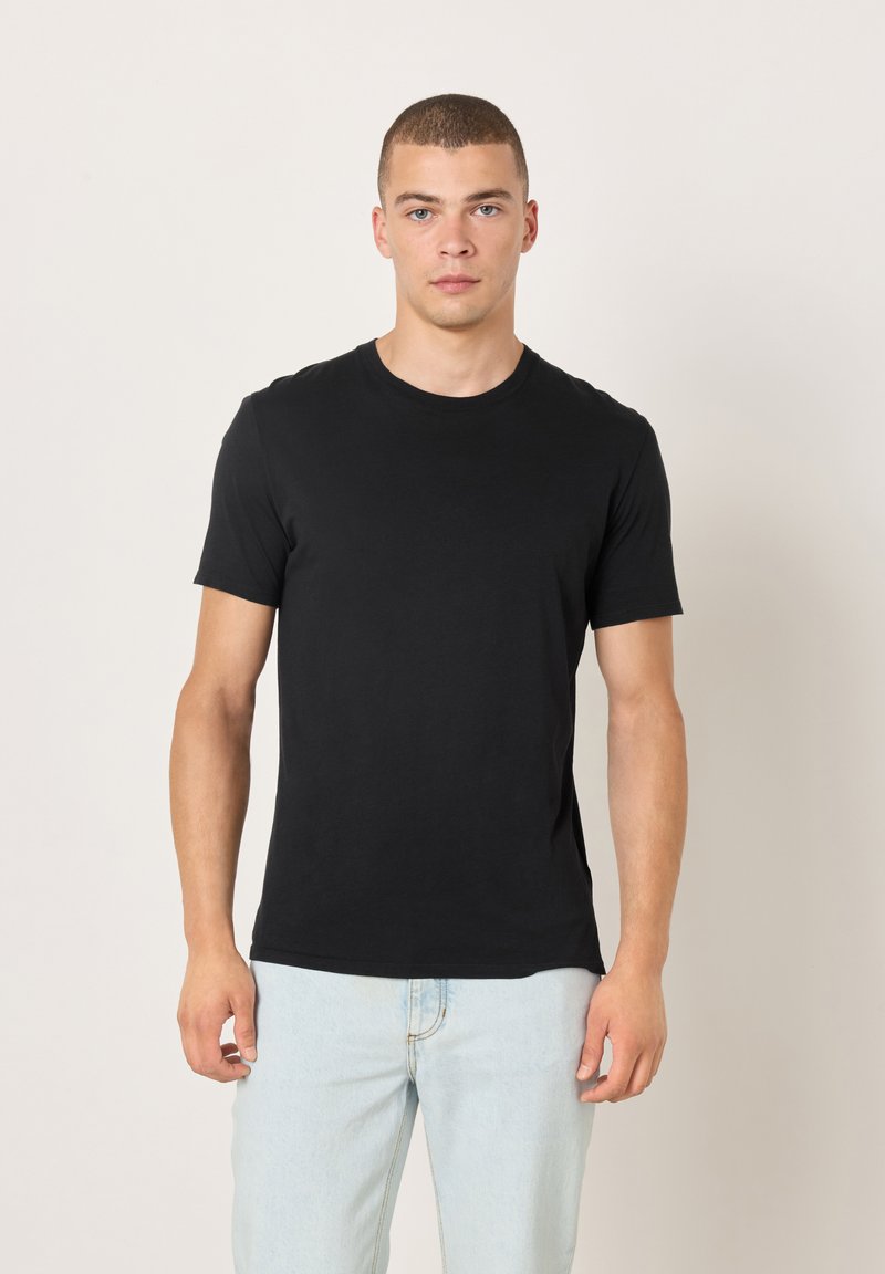 Crna pamučna majica s okruglim izrezom, kratkim rukavima i prilagođenim krojem. Nošena s svijetloplavim trapericama, prikazana na neutralnoj pozadini.