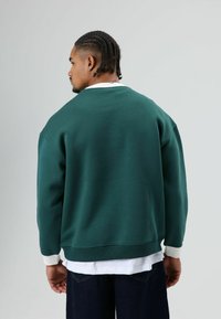 Groen oversized sweatshirt met geribbelde boorden, ronde halslijn en een gelaagd wit overhemd eronder. Casual pasvorm, zachte textuur, minimalistisch ontwerp.
