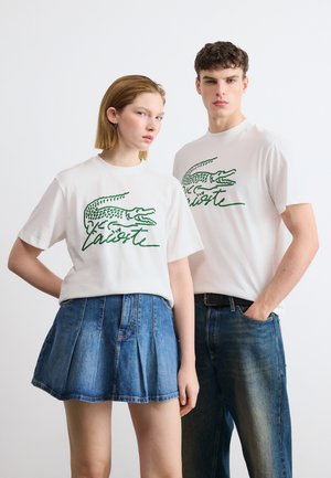 Dwie modelki w białych koszulkach z logo Lacoste; jedna zestawiła je z dżinsową spódnicą, druga z niebieskimi dżinsami i czarnym paskiem.