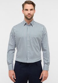 Man in een wit en donkergroen gestreept overhemd met lange mouwen en een broek in een donkere kleur, staand tegen een effen witte achtergrond.