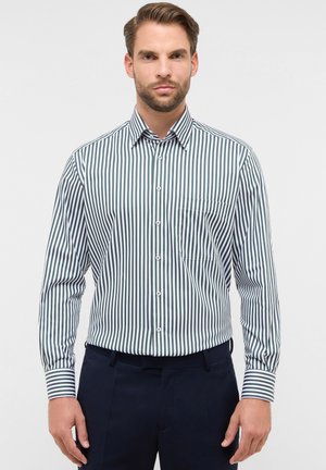Man in een wit en donkergroen gestreept overhemd met lange mouwen en een broek in een donkere kleur, staand tegen een effen witte achtergrond.