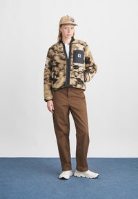Chaqueta de polar con un patrón en tonos marrones y negros, con cremallera frontal y bolsillo en el pecho; combinada con pantalones marrones y zapatillas blancas, bajo una gorra beige.