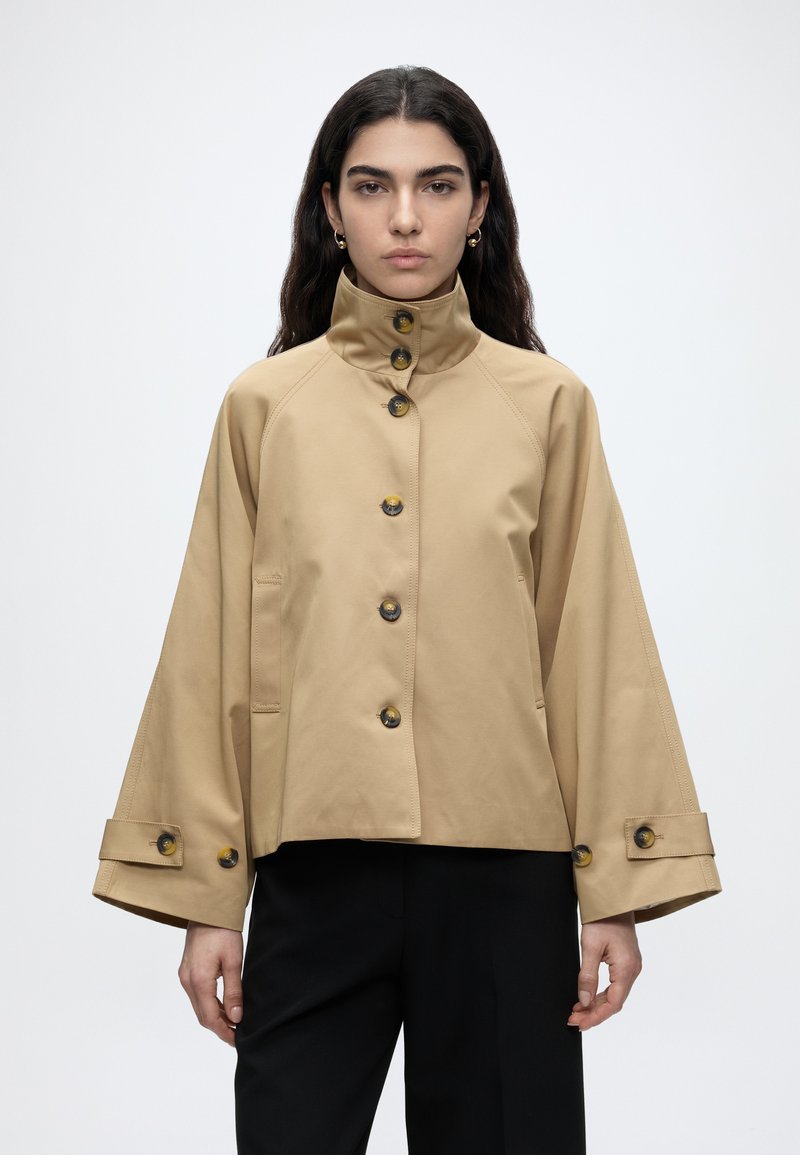 Femme aux longs cheveux foncés portant une veste beige à col montant boutonnée avec des manches larges et un pantalon noir, debout devant un fond uni.