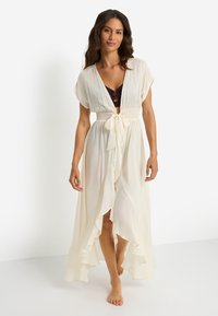 Μακρύ, αέρινο κάλυμμα παραλίας σε εκρού σιφόν· διαθέτει βαθύ V-λαιμόκοψη, κοντά μανίκια, κορδόνι στη μέση και βολάν στο τελείωμα με μαλακή, ελαφριά υφή.