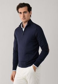 Muž v tmavě modrém svetr s půlzipem přes světle modrou košili a bílé kalhoty, stojící s jednou rukou v kapse před jednobarevným pozadím.