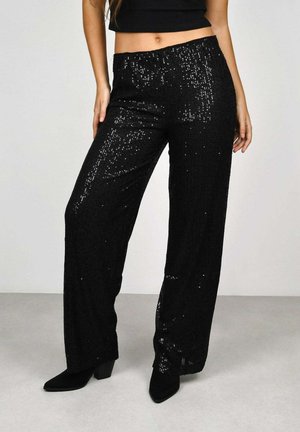 Mujer con pantalones anchos de lentejuelas negras, botas negras de punta puntiaguda hasta el tobillo y un top negro corto, de pie sobre un suelo gris claro.