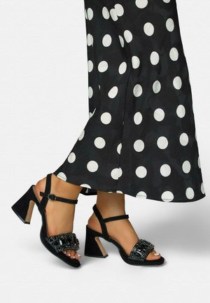 Sandalias negras de tacón ancho con tiras en el tobillo y bandas delanteras adornadas con joyas, combinadas con pantalones anchos negros con lunares.