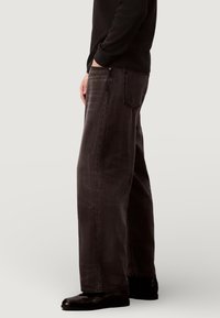 Jean en denim gris foncé à coupe décontractée, avec des détails de couture remarquables, des poches latérales et une ceinture standard, porté avec des chaussures noires.