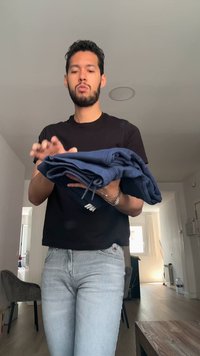 Άνδρας με μαύρο πουκάμισο και ανοιχτόχρωμα τζιν διπλώνει ή κρατάει ένα ζευγάρι μπλε φούτερ μέσα σε μοντέρνο διαμέρισμα.