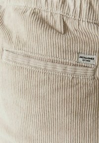 Beige kordbársony anyag függőleges bordákkal és vízszintes zsebvarrással, amelyen egy kis fehér Jack & Jones címke fekete szöveggel.