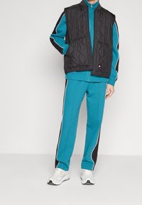 Svart vadderad väst över en turkos zip-up jacka och matchande sweatpants med svarta sidpanelen och vita ränder. Vita sneakers fulländar looken.