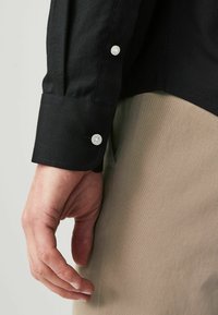 Schwarzes Longsleeve-Shirt aus strukturiertem Material mit einem gewellten Ärmel und zwei weißen Knöpfen. Die Hand ruht auf einem hellen khakifarbenen Stoff.