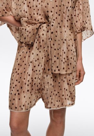 Pantalones cortos de tela ligera marrón con lunares irregulares más oscuros combinados con una camisa suelta a juego, mano en la cadera, fondo blanco.