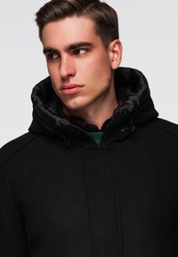 Černý kabát z vlněné směsi s vysokým límcem a odnímatelnou saténovou kapucí; má zipové zapínání a minimalistické designové prvky.