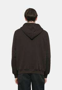 Felpa marrone con cappuccio dal taglio ampio e rilassato, caratterizzata da una texture liscia, polsini a costine e un design semplice senza loghi o motivi visibili.