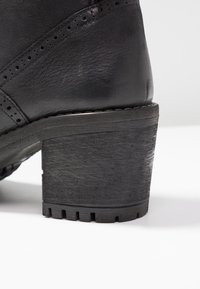 Stivale in pelle nera con tacco dal design impilato testurizzato e dettagli perforati vicino al bordo superiore su sfondo bianco.