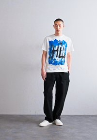 Weißes T-Shirt mit einem abstrakten blauen und schwarzen Grafikdesign. Kombiniert mit lockeren schwarzen Hosen und weißen Sneakers. Schlichte, moderne Silhouette.