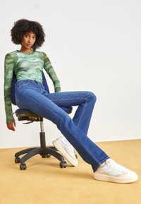 Pantalones de mezclilla acampanados azules con costuras visibles, combinados con una camisa de manga larga verde con un patrón ondulado y zapatillas deportivas blancas.