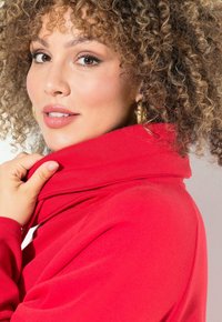 Maglione rosso con colletto alto, tessuto testurizzato e vestibilità rilassata. Il modello ha i capelli ricci e indossa orecchini a cerchio dorati, mentre tiene il colletto.