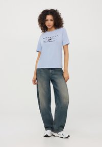 Jasnoniebieska bawełniana koszulka z czarnym nadrukiem, w połączeniu z luźnymi, wyblakłymi niebieskimi dżinsami i białymi sneakersami. Widoczne kręcone włosy.