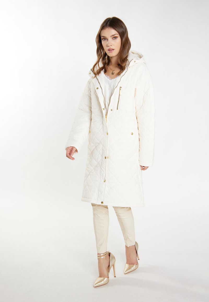 Gequilte witte lange jas met een capuchon, gouden hardware en een rits aan de voorkant. Draagt over crèmekleurige slim-fit broeken en gouden hoge hakken. Soepele textuur.