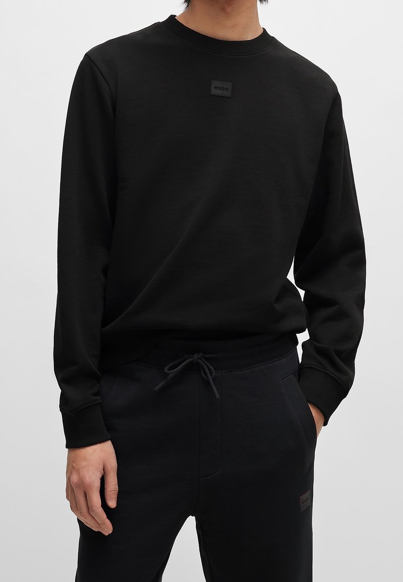Personne portant un sweat-shirt noir à manches longues et un pantalon noir à cordon avec un petit patch rectangulaire « Hugo » sur la poitrine.