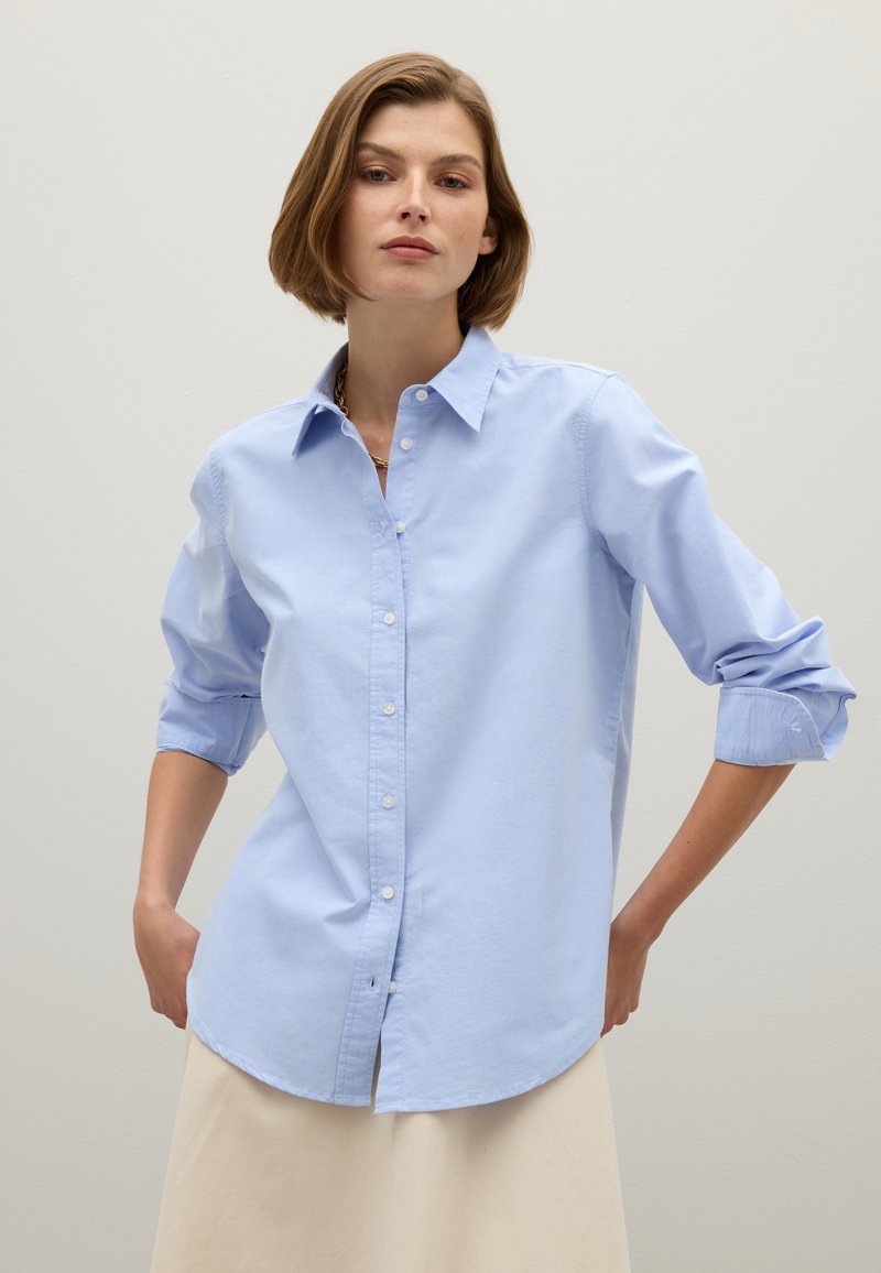 Light blue button-up shirt with long sleeves rolled to the elbows, featuring a classic collar and a curved hem. Smooth, cotton texture.