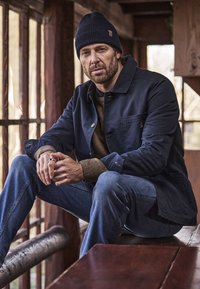 Jakna tamnoplave boje s dugmadima, bež pleteni džemper, plave traperice i tamnoplava kapa; sjedi u zatvorenom prostoru s drvenim detaljima u pozadini.