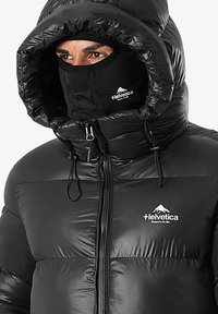 Personne portant une veste rembourrée noire avec capuche et un masque facial noir assorti, tous deux arborant un logo "Helvetica" blanc avec un design de montagne.