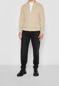 Mężczyzna w beżowym swetrze na zamek, białej koszuli, czarnych dżinsach i czarnych skórzanych butach, stojący na tle gładkiego białego tła.