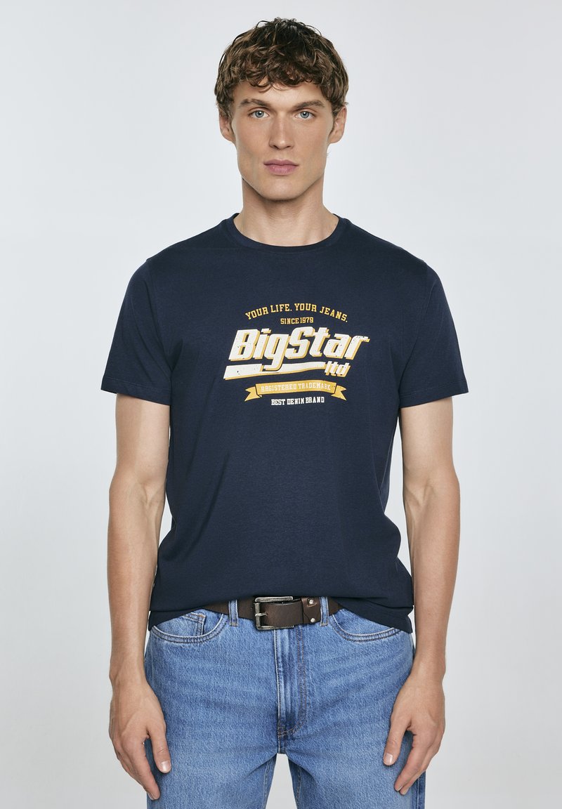 Jūros spalvos marškinėliai su apvaliu kaklu, su baltais ir geltonais grafikos užrašais. Dėvimi su mėlynomis džinsinėmis kelnėmis ir ruduo diržu. Lygus medvilnės tekstūras.