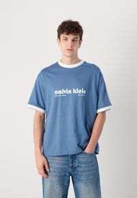 Blue cotton t-shirt with white ribbed collar and sleeve cuffs. Features bold "calvin klein" logo text and New York location details.