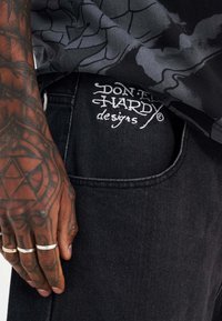 Black denim jeans with embroidered "Don Hardy designs" logo on the back pocket. Skin texture visible on a hand with multiple rings.