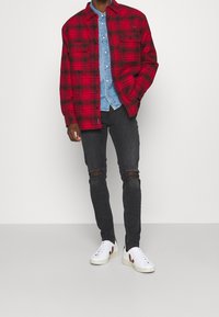 Camisa de franela a cuadros rojos y negros sobre una camisa de mezclilla azul clara, combinada con jeans negros desgastados y zapatillas blancas con acentos rojos.