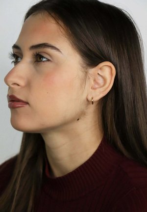 Jeune femme aux longs cheveux bruns portant un col roulé bordeaux et une petite boucle d'oreille en or, regardant vers la gauche sur un fond uni.
