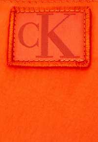 Pomarańczowa metka materiałowa z obszytym brzegiem, z przezroczystym tłem i literami „CK” w ciemniejszym odcieniu pomarańczy.