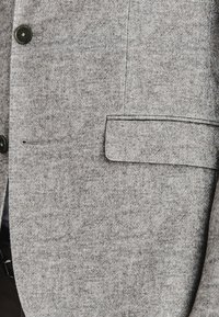 Szary, wełniany żakiet z delikatną teksturą, dwa guziki i klapami typu notch. Zawiera kieszeń na piersi oraz detale przy szwach bocznych.