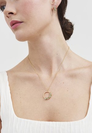 Kobieta nosząca złoty naszyjnik w kształcie kwiatowego koła i pasujące kolczyki, ubrana na biało, z włosami związanymi w kok na gładkim tle.