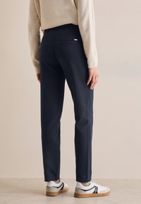 Navyblauwe tailored broek met een gladde textuur, voorzien van achterzakken en een slim-fit ontwerp. Gepaard met witte sneakers.