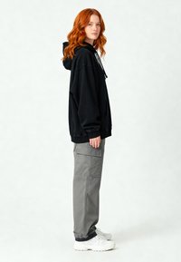 Giovane con lunghi capelli rossi, che indossa un felpa nera con cappuccio, pantaloni cargo grigi e sneaker bianche, in piedi di lato su uno sfondo semplice.